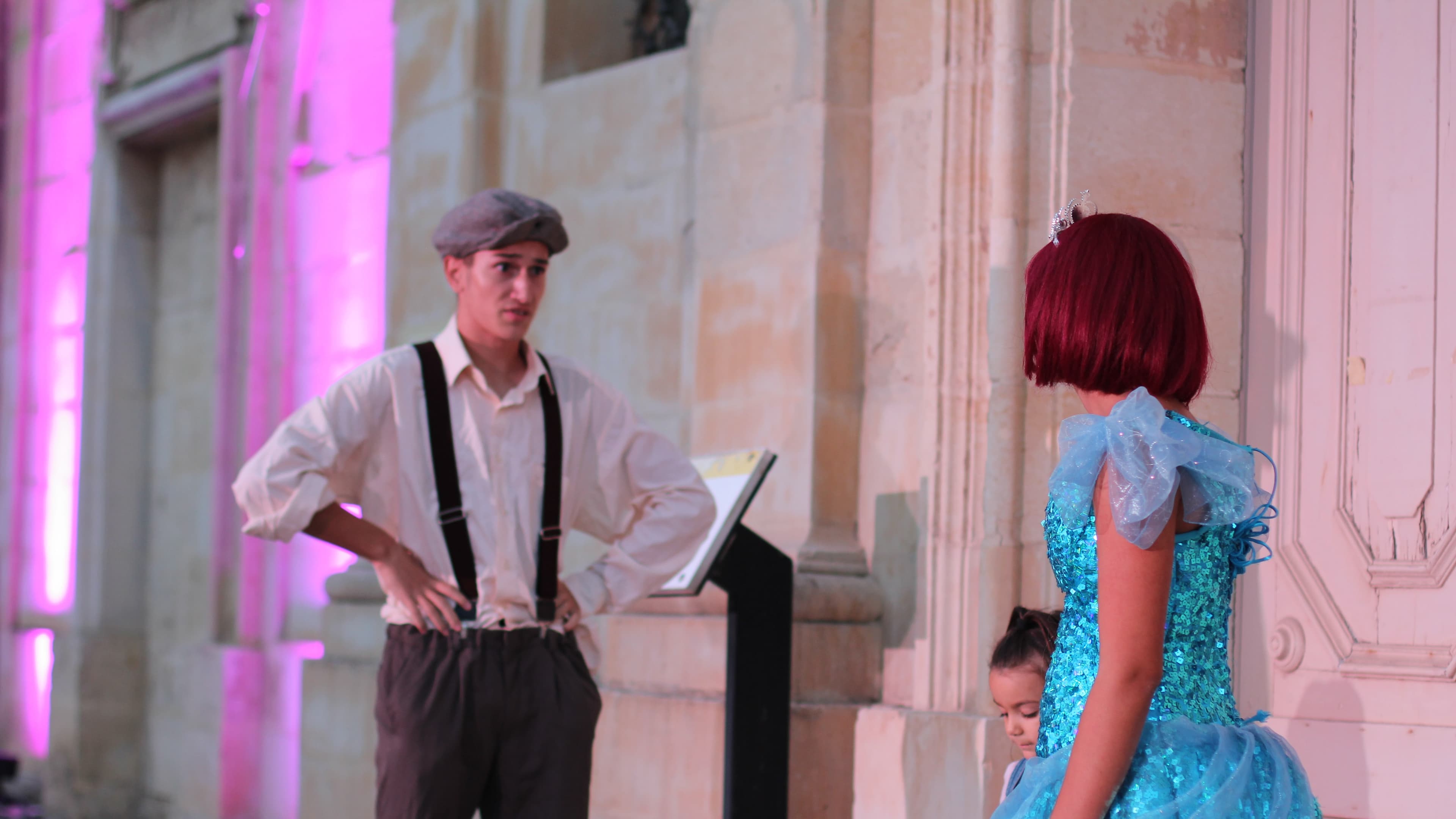 A performer in a cap and suspenders faces a girl in a sequinned turquoise dress on a stone-walled stage